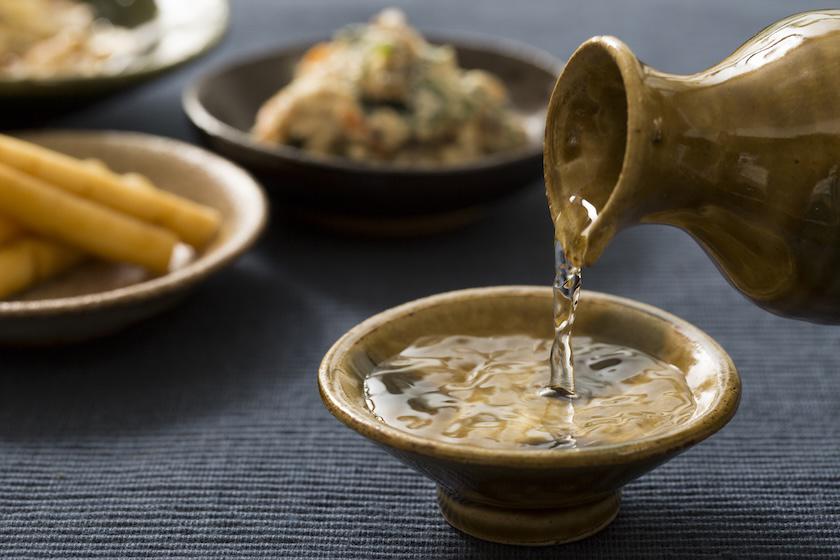 背景には料理が盛られた小皿が並び、おちょこに日本酒を注いでいる。