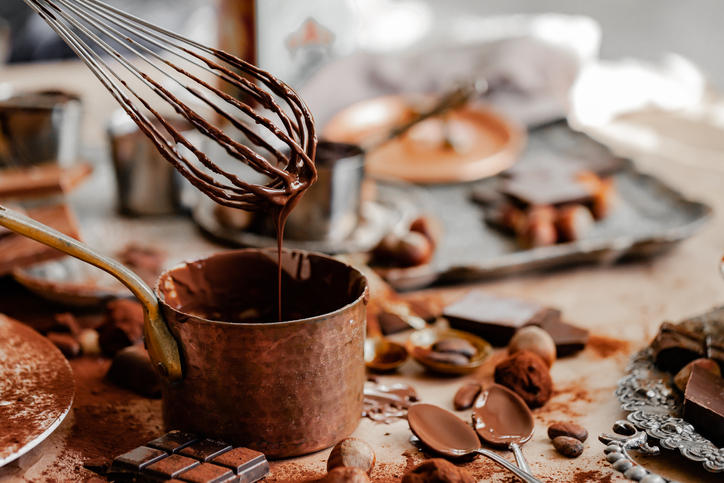 キッチンの上に広げられた食器や調理器具、チョコレートと、銅の片手鍋で溶かされたチョコレートを泡立て器で持ち上げている場面