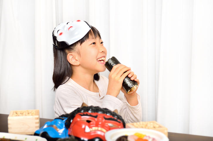 女の子がお面をかぶりながら恵方巻きを食べようとしている