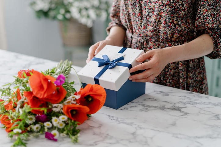 オレンジの花で作られた小さいブーケと青いリボンがかかった白い正方形のプレゼントボックスを開けようとする女性の手