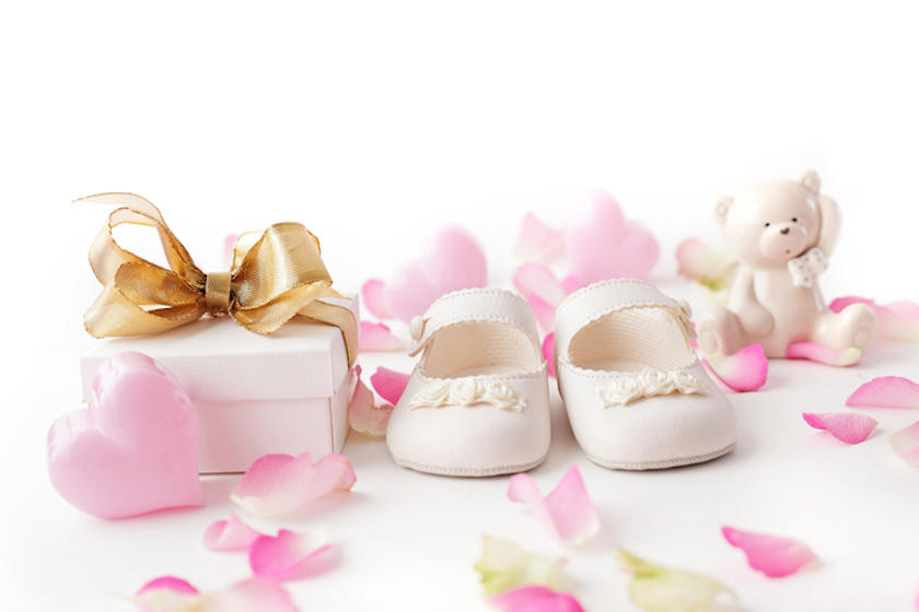 赤ちゃん用の白い靴と、金色のリボンがかかった白いギフトボックスが並び、周りにピンク色の花びらが散っている。