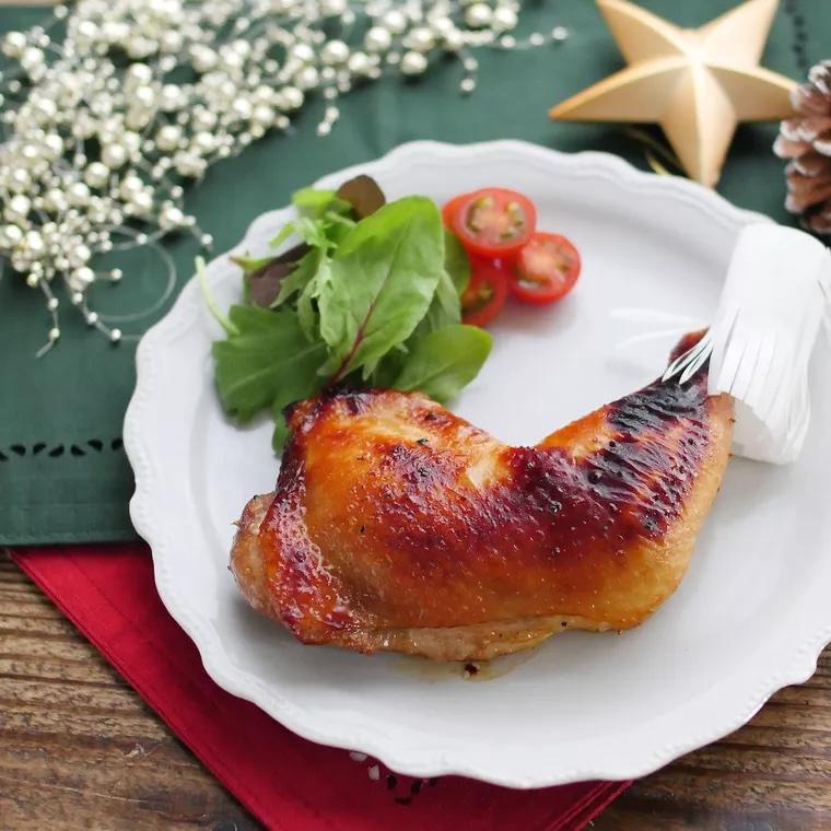 クリスマスのテーブルクロスの上に、鶏もも肉のローストチキンがのった白い皿が置かれている。