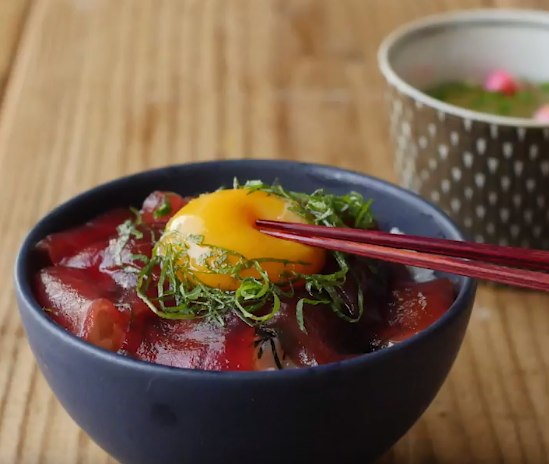 木製テーブルの上にのっているトッピングの卵黄に箸が添えられたまぐろの漬け丼が