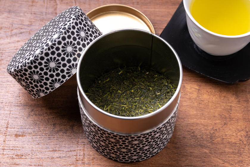 緑茶の茶葉入りの缶がテーブル中央に置かれ、右上に緑茶が入った湯呑みが置かれている。