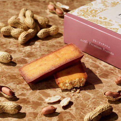 ダイスカットのピーナッツが入ったフィナンシェ2個と、ピンク色の箱が、茶色いテーブルの上に置かれており、そのまわりには落花生が散りばめられている