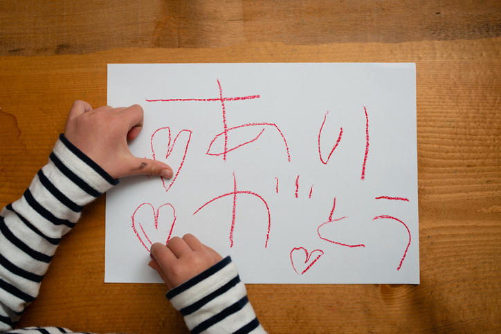 画用紙に赤いクレヨンで「ありがとう」と書く子どもの手もと
