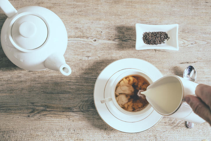 木のテーブルにミルクを入れている紅茶入りティーカップとティーポットと白いお皿にのせられた茶葉が置かれている