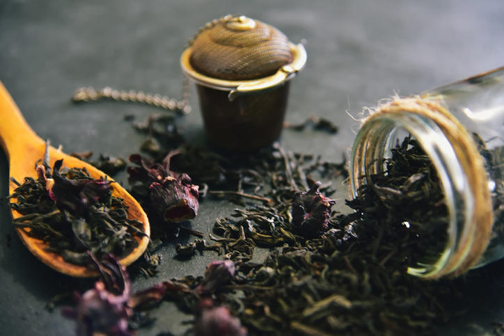 紅茶の茶葉がのった木のスプーンが、黒いテーブルの上に置かれており、その横には倒れたガラス瓶からあふれ出た紅茶の茶葉が散らばっている