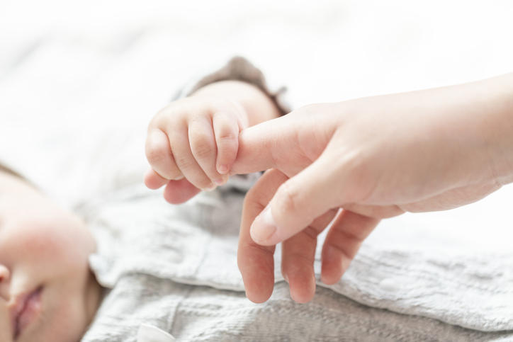 白い肌着を着た赤ちゃんが大人の指を握っている