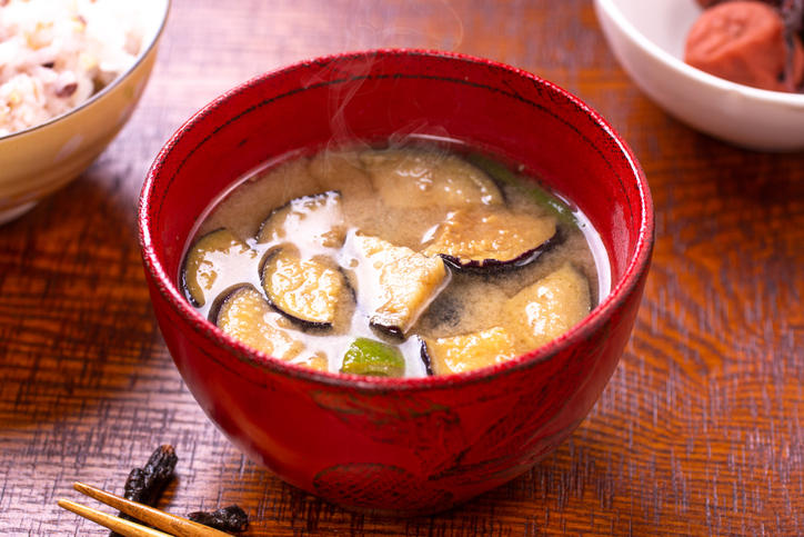 テーブルの上の赤い汁椀に入ったなすの味噌汁、背景に雑穀ご飯と梅干し