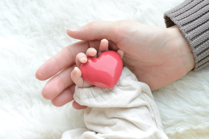 赤いハートの小物を持っている赤ちゃんの手を、大人の手が包み込んでいる写真