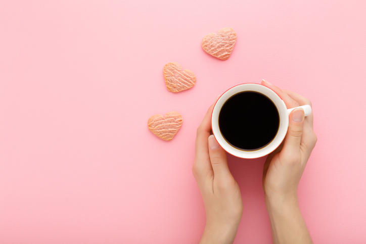 ピンク色のテーブルに置かれた白いカップ入りのコーヒーに両手を添えている様子
