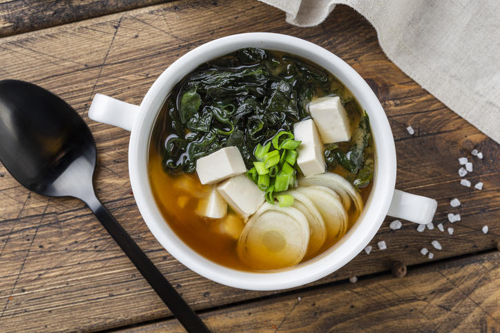 木のテーブルの上の白いボウルに入ったわかめ、豆腐、ねぎの味噌汁と黒いスプーン