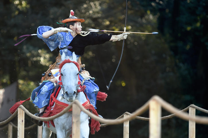 女性が白い馬に乗りながら流鏑馬をしている