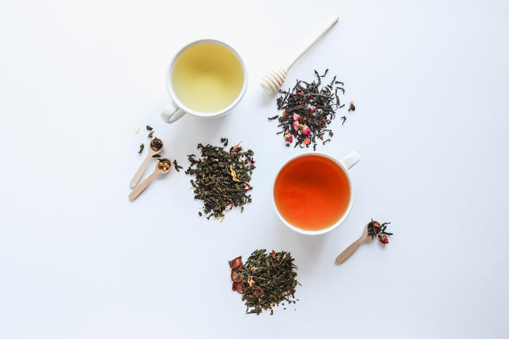 白いテーブルの上に紅茶や緑茶の茶葉とお茶が注がれた白いカップと茶葉が乗せられた木のスプーンが置かれている