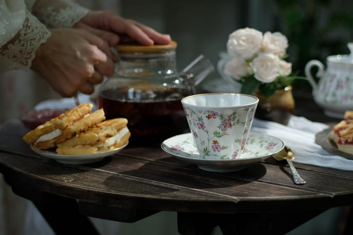 木のテーブルに花柄のティーカップと紅茶が入ったガラスポットとスイーツ、花が置かれている
